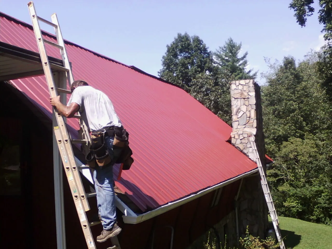 Roofer on a ladder beside a red metal roof and stone chimney — Soffit & Fascia Repair by Roofing Brothers in Port Arthur
